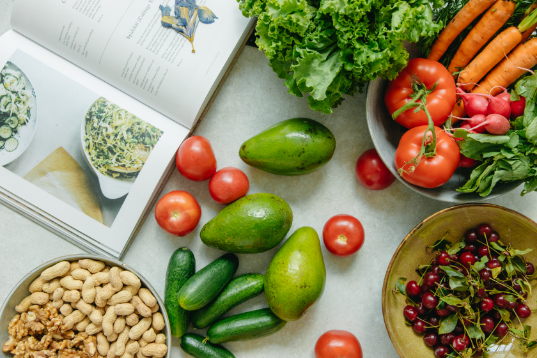 Assorted fresh vegetables and fruits, including avocados, cucumbers, tomatoes, radishes, carrots, cherries, and peanuts, displayed around an open cookbook on a marble surface.